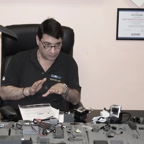 Francesco Polimeni, fondatore di Polinet S.r.l. ed ex agente della Polizia di Stato, durante una sessione formativa del corso TSCM a Roma. Il docente spiega le caratteristiche tecniche di microspie, microtelecamere e dispositivi di intercettazione disposti sulla scrivania, con un certificato di specializzazione visibile sullo sfondo.