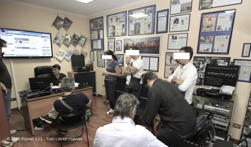 Sessione pratica del corso TSCM di Polinet S.r.l. a Roma con il docente che illustra l'utilizzo della strumentazione professionale per la bonifica ambientale. I corsisti, con volti oscurati per privacy, osservano attentamente le dimostrazioni. Sullo sfondo le pareti dell'aula decorate con certificazioni e rassegna stampa, e le valigette con l'attrezzatura tecnica per la controsorveglianza.